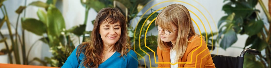 Collaborative discussion in an office setting; two individuals seated with a tablet, surrounded by indoor plants.