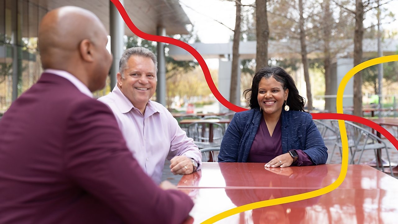 Group discussion in an outdoor setting, featuring two individuals in a white shirt and blue top, with a blurred city park backdrop
