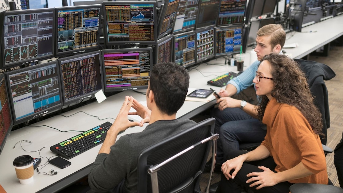 A group of colleagues gathered around multiple trading monitors displaying financial data in a busy workspace.