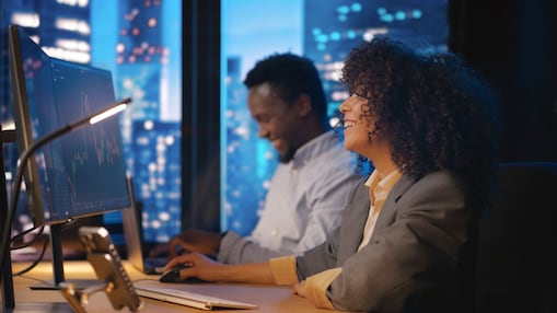 Two people working at computer stations in an office at night, with financial charts displayed on a large monitor.