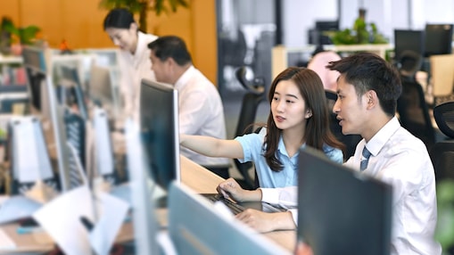 Office workers collaborating at their desks, with one person pointing at a computer screen.