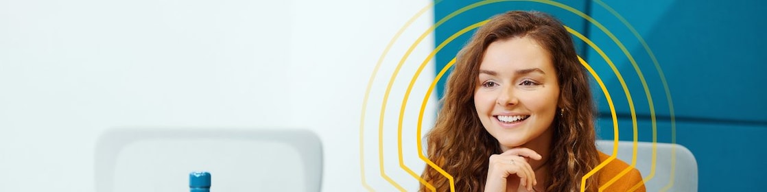 A person with long curly hair, wearing an orange sweater, is sitting at a white table in front of an orange laptop. There is a blue water bottle on the table to the left of the laptop.
