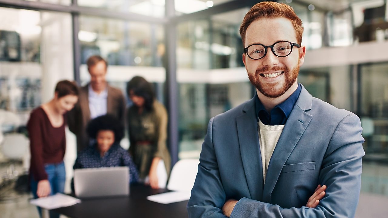 Portrait of a confident young man standing in a modern office with his colleagues in the background