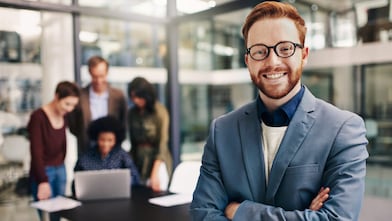 Portrait of a confident young man standing in a modern office with his colleagues in the background