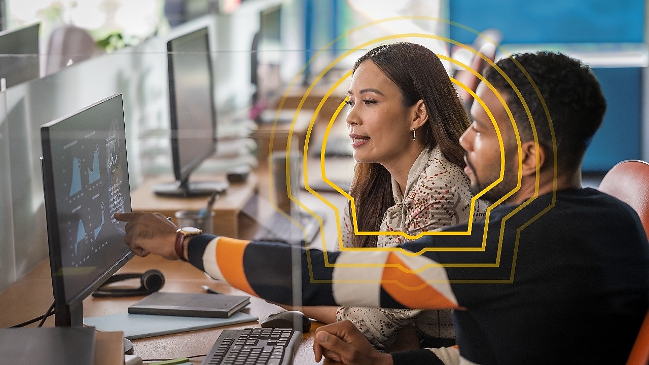 Two people working at a desk, viewing data charts on a computer monitor in a modern office setting.