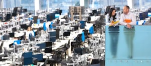 Open-plan office with desks and monitors; two people on a balcony talking, overlooking a busy workspace.