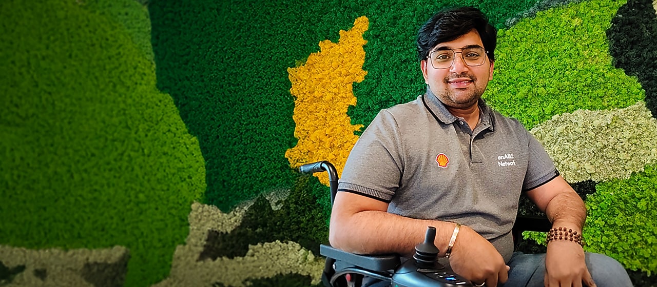 Person in a wheelchair wearing a Shell enABLE Network shirt, seated against a green and yellow moss wall.