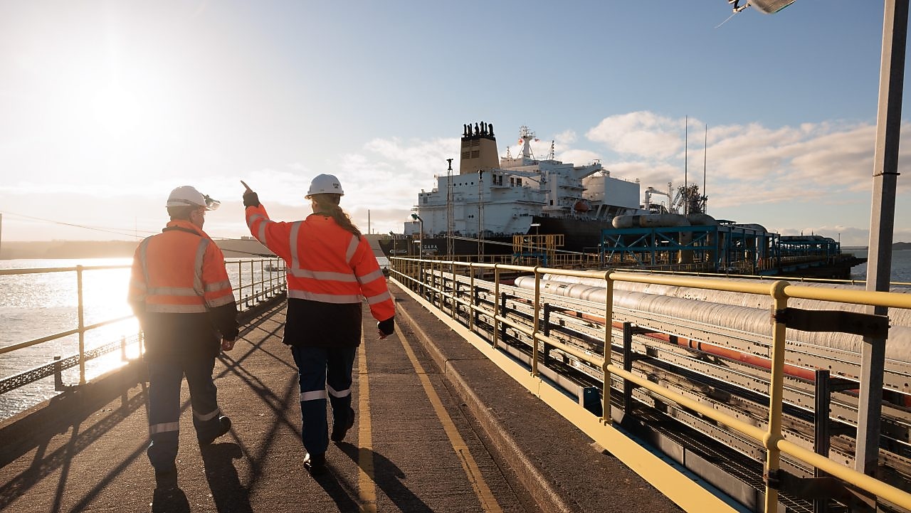 People walking along a dock with industrial structures in the background.