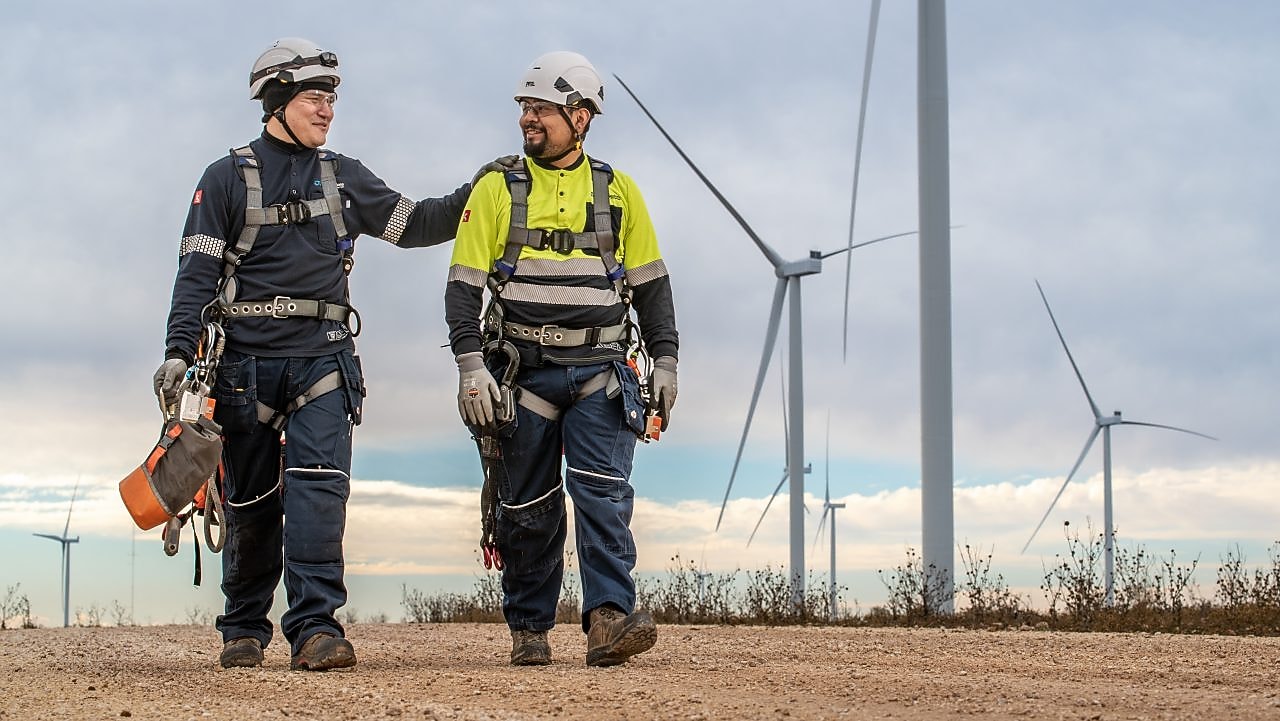 Two individuals wearing safety gear standing near wind turbines.