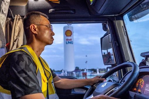 A truck driver wearing a yellow safety vest seated inside a truck cab, holding the steering wheel, with a Shell LNG fuelling station visible through the windshield..