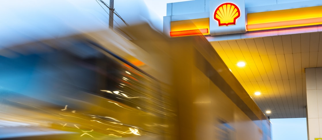 A yellow express delivery truck in motion at a Shell fuelling station, with the Shell logo visible on the canopy.