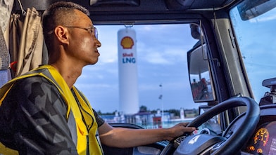 A truck driver wearing a yellow safety vest seated inside a truck cab, holding the steering wheel, with a Shell LNG fuelling station visible through the windshield.