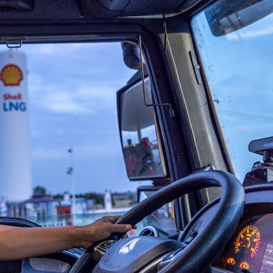 A truck driver wearing a yellow safety vest seated inside a truck cab, holding the steering wheel, with a Shell LNG fuelling station visible through the windshield.