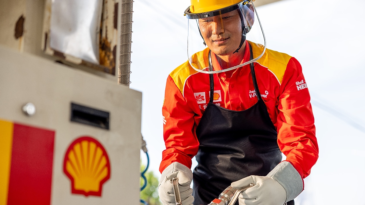 A Shell station worker wearing a red and yellow uniform, safety helmet, and gloves, connecting a fuel hose to equipment at an LNG fuelling station.