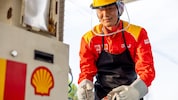A Shell station worker wearing a red and yellow uniform, safety helmet, and gloves, connecting a fuel hose to equipment at an LNG fuelling station.