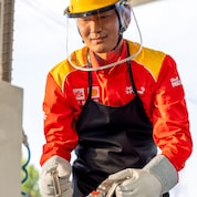 A Shell station worker wearing a red and yellow uniform, safety helmet, and gloves, connecting a fuel hose to equipment at an LNG fuelling station.