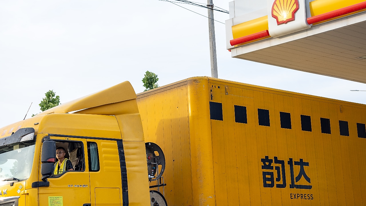 A yellow express delivery truck parked at a Shell fuelling station, with the Shell logo visible on the canopy and a driver seated inside the cab.
