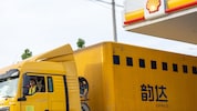 A yellow express delivery truck parked at a Shell fuelling station, with the Shell logo visible on the canopy and a driver seated inside the cab.