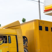A yellow express delivery truck parked at a Shell fuelling station, with the Shell logo visible on the canopy and a driver seated inside the cab.