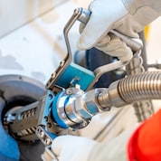 Close-up of a Shell station worker wearing gloves and a red uniform, removing an LNG fuelling nozzle from a dispenser.