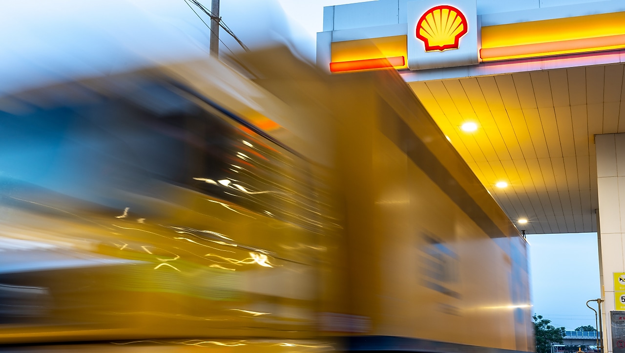 A yellow express delivery truck in motion at a Shell fuelling station, with the Shell logo visible on the canopy.