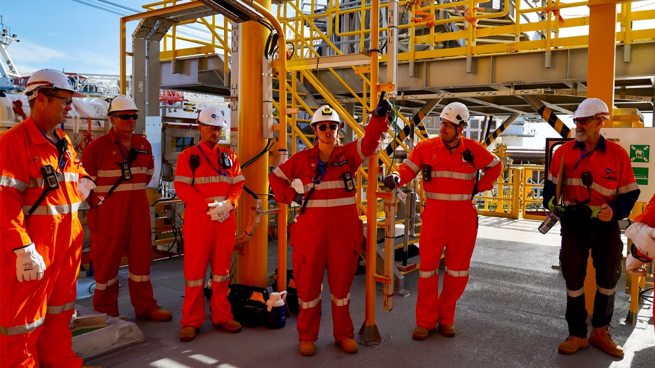 Emma stands with members of the offshore team on the Crux facility, wearing safety coveralls and hard hats among industrial equipment and walkways.​