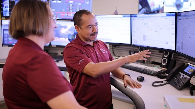 Two Shell engineers, their t-shirts maroon with a white whale logo, stand in front of the computer screens in a control room in New Orleans. 