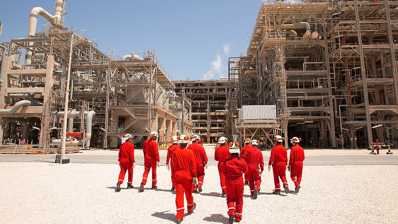 A cluster of around a dozen workers in red overalls and white hard hats walks towards a large energy facility.