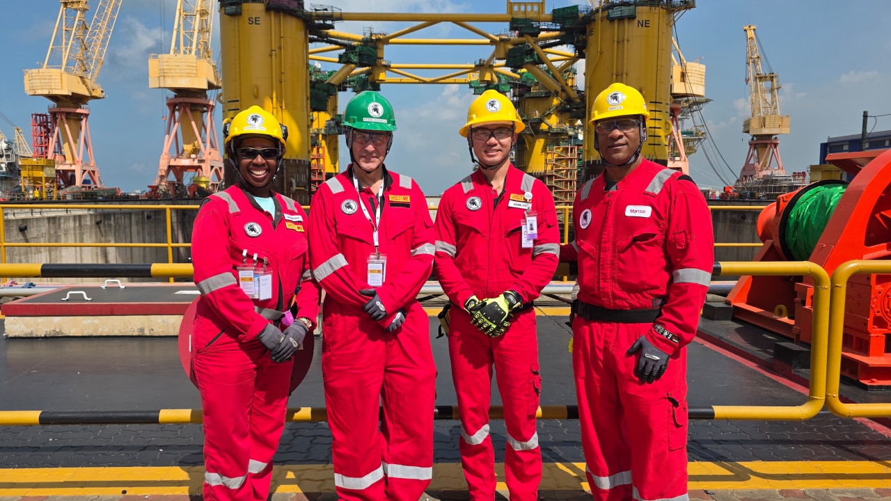 Four workers in red coveralls, gloves, and yellow safety helmets stand side by side on a dock in front a large yellow hull of the offshore platform under construction in the background. Cranes, scaffolding, safety railings, cables, and heavy machinery are visible around them under a clear sky.