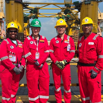 Four workers in red coveralls, gloves, and yellow safety helmets stand side by side on a dock in front a large yellow hull of the offshore platform under construction in the background. Cranes, scaffolding, safety railings, cables, and heavy machinery are visible around them under a clear sky.