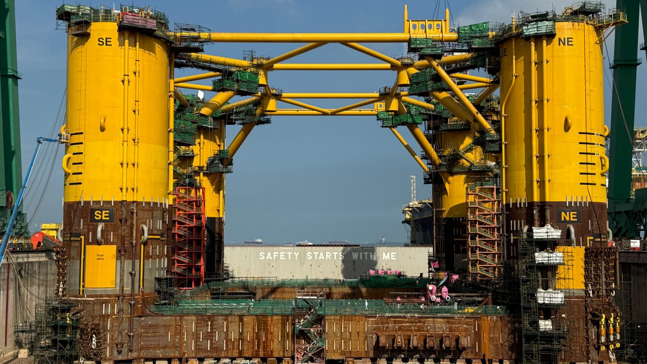 Large yellow offshore platform hull under construction in a dry dock in Singapore, with four tall cylindrical columns connected by steel beams and surrounded by scaffolding, cranes, and construction equipment. Workers, vehicles, and materials are visible at ground level, highlighting the scale of the structure.