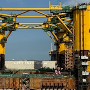 Large yellow offshore platform hull under construction in a dry dock in Singapore, with four tall cylindrical columns connected by steel beams and surrounded by scaffolding, cranes, and construction equipment. Workers, vehicles, and materials are visible at ground level, highlighting the scale of the structure.