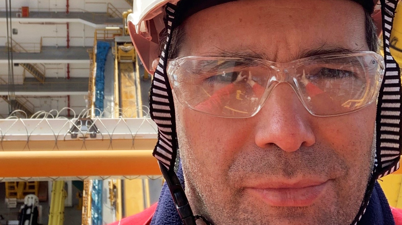 Close-up of a worker wearing a white safety helmet with a Shell logo and clear protective glasses, standing on a drillship. In the background, the vessel’s deck is visible with a circular helipad, tower structure, and equipment, with calm sea and cranes and ships in the distance.