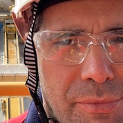 Close-up of a worker wearing a white safety helmet with a Shell logo and clear protective glasses, standing on a drillship. In the background, the vessel’s deck is visible with a circular helipad, tower structure, and equipment, with calm sea and cranes and ships in the distance.