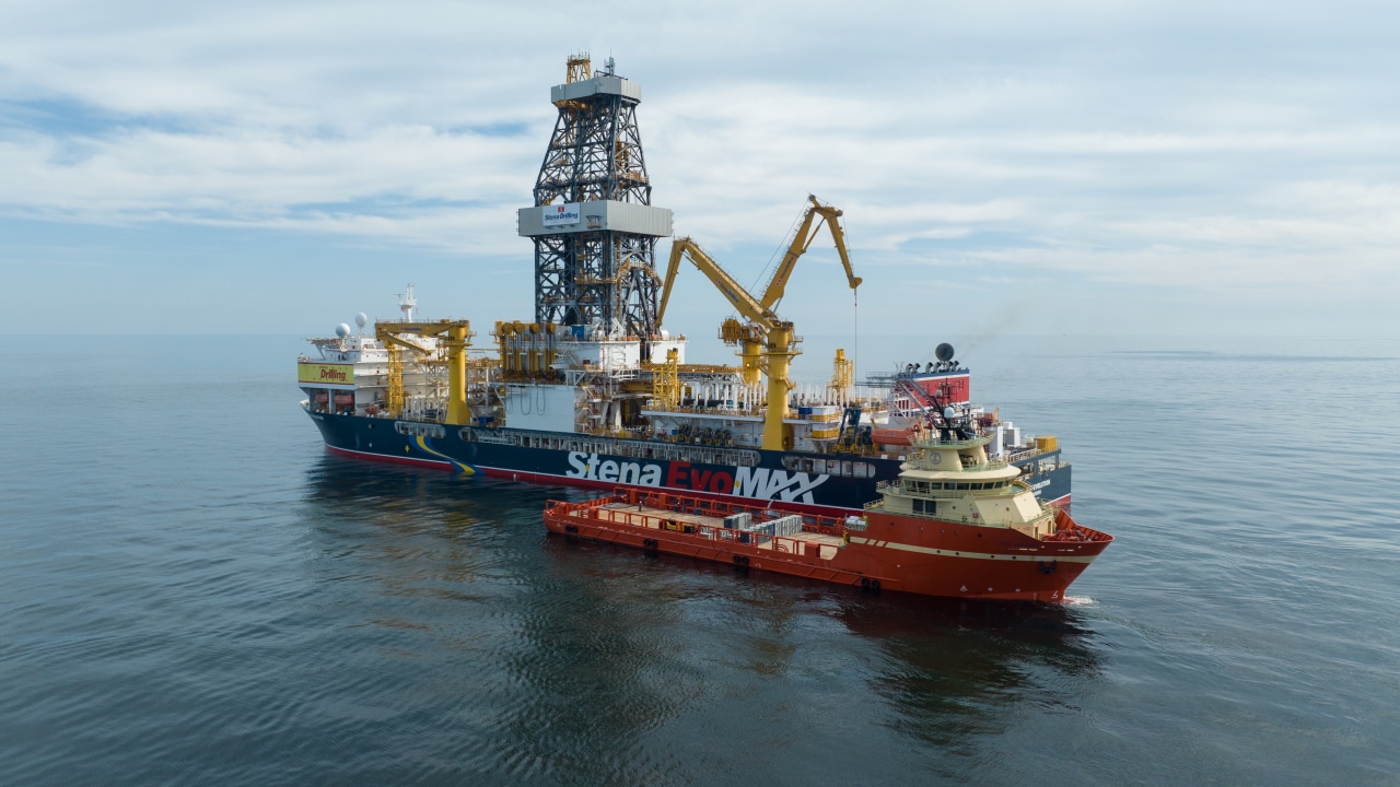 A large offshore drillship sails on calm open water, featuring a tall central drilling derrick and multiple yellow cranes. A smaller red support vessel is positioned alongside the drillship, with equipment visible on its deck under a cloudy sky.