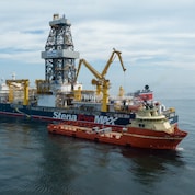 A large offshore drillship sails on calm open water, featuring a tall central drilling derrick and multiple yellow cranes. A smaller red support vessel is positioned alongside the drillship, with equipment visible on its deck under a cloudy sky.