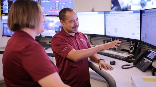 Two Shell engineers, their t-shirts maroon with a white whale logo, stand in front of the computer screens in a control room in New Orleans. 
