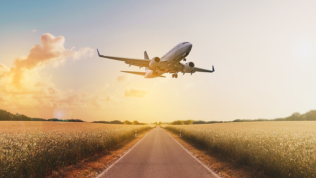 Aeroplane taking off over a road surrounded by fields at sunrise.