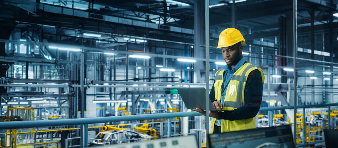A male engineer wearing a yellow hard hat and high-visibility vest stands in a modern, high-tech automotive factory. 