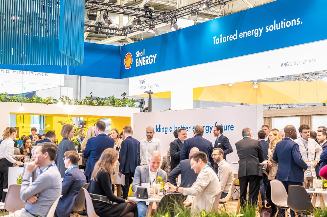 A light and open Shell Energy event stand in an exhibition hall with the Shell Energy logo at the top of the stand. Many men and women standing and seated at tables having business conversations.