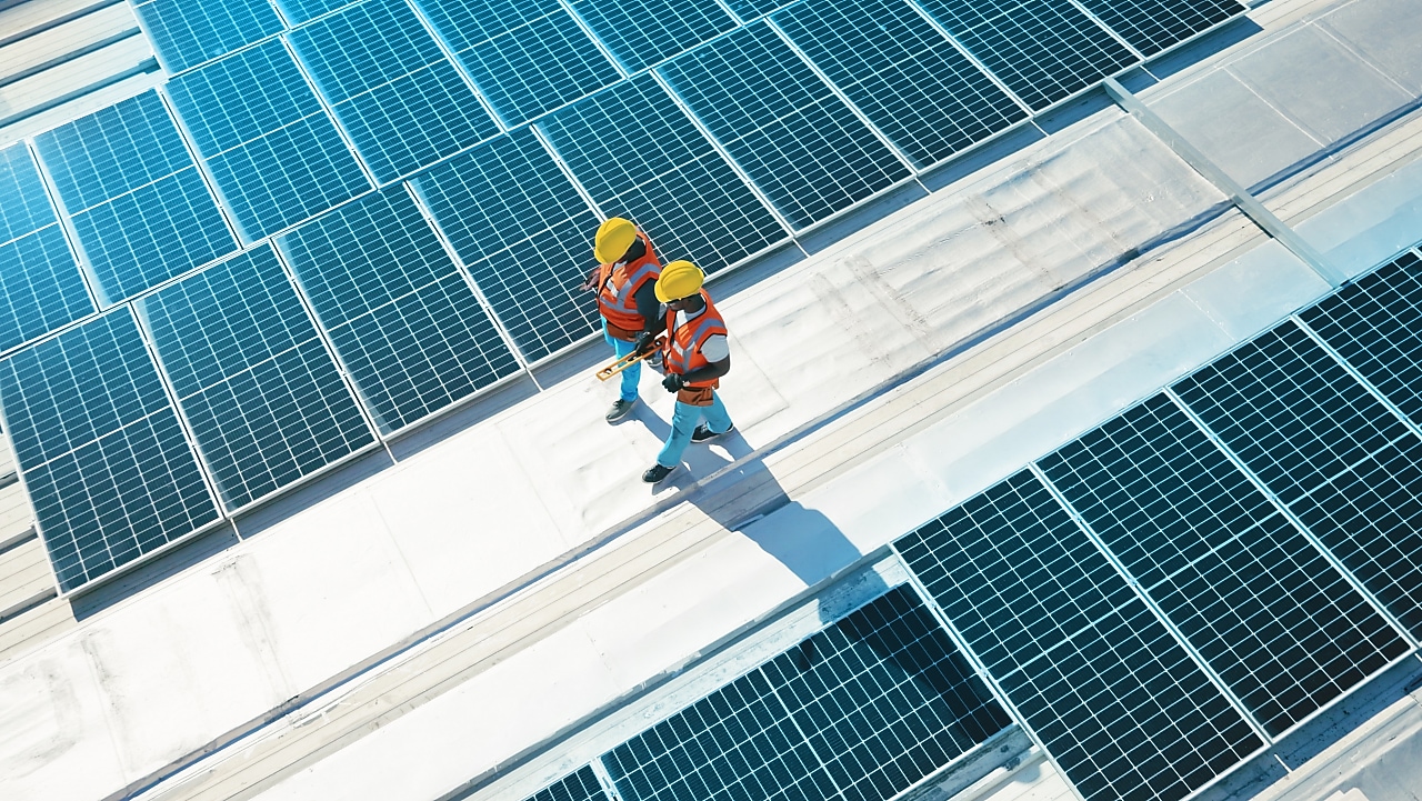 Two workers wearing hard hats and safety vests walk across a rooftop covered with solar panels, inspecting the installation under bright sunlight.