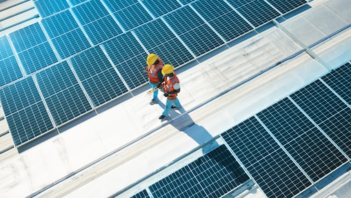 Two workers wearing hard hats and safety vests walk across a rooftop covered with solar panels, inspecting the installation under bright sunlight.