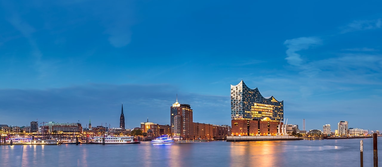 Panoramic evening view of Hamburg’s waterfront, featuring the illuminated Elbphilharmonie concert hall rising above the harbor, with boats docked along the water and the city skyline under a clear blue sky.