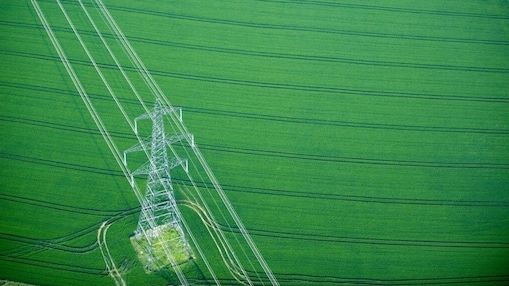 Aerial view of a green agricultural field with a tall electricity pylon and multiple power lines running diagonally across the image.