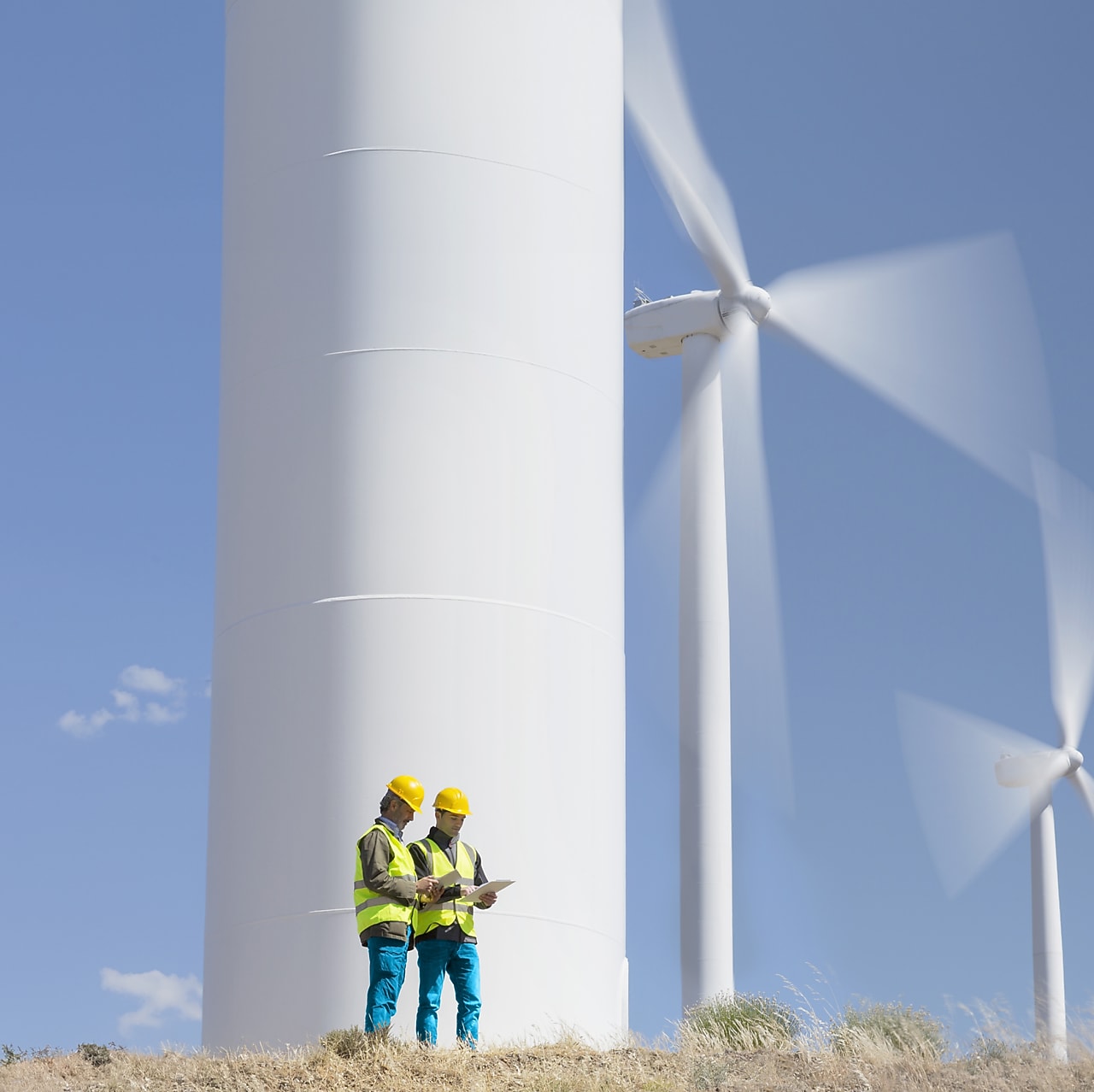 Two engineers wearing safety gear stand at the base of a large wind turbine, reviewing documents, with additional wind turbines spinning in the background against a clear blue sky.