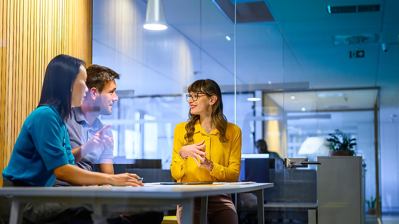 Three colleagues sit around a table in a modern glass-walled office, engaged in a friendly conversation, with warm lighting and contemporary workspace elements in the background.