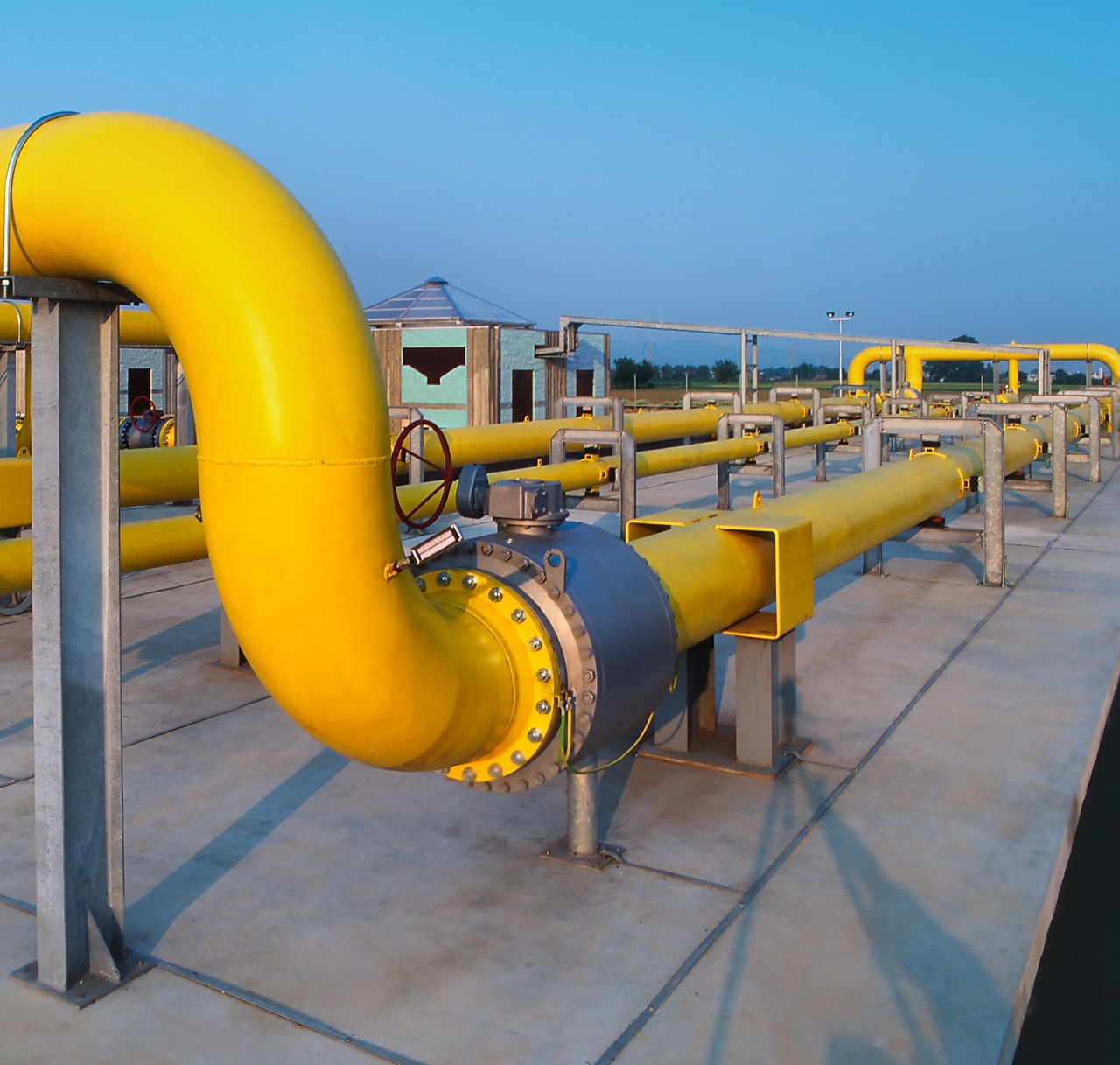 A network of large yellow industrial pipelines installed on a concrete platform at an outdoor facility, with valves and support structures visible under clear blue sky.