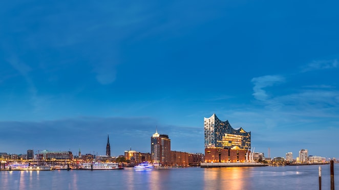Panoramic evening view of Hamburg’s waterfront, featuring the illuminated Elbphilharmonie concert hall rising above the harbor, with boats docked along the water and the city skyline under a clear blue sky.