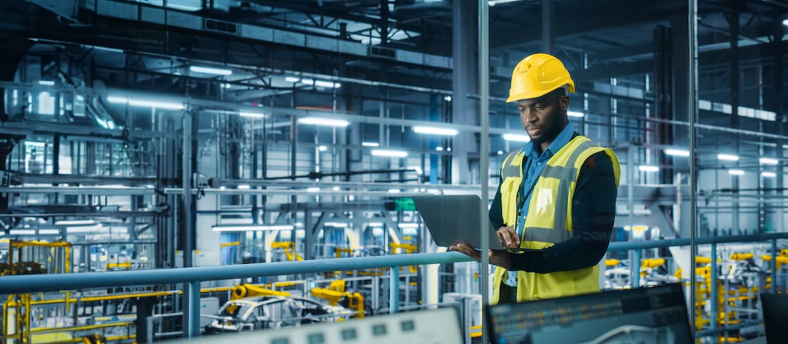 A male engineer wearing a yellow hard hat and high-visibility vest stands in a modern, high-tech automotive factory. 