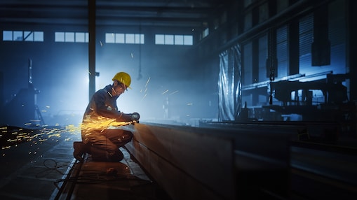 Industrial worker wearing safety gear, including a yellow helmet and protective glasses, kneels while using a power tool to cut a metal beam in a dimly lit factory.